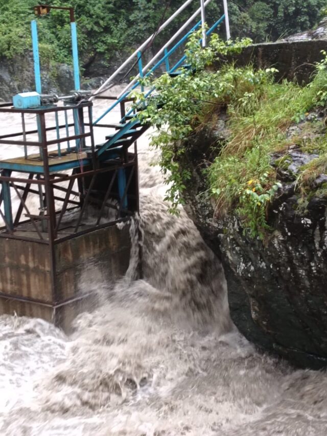 A strong flow of water rushing through a metal water control structure with a staircase, surrounded by greenery and rocks.